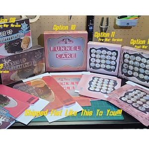 May include: A collection of eight different box designs for a board game. The boxes are all different colors and have different designs. The boxes are all flat and ready to be shipped. The text on the boxes reads "Saddle Up Salisbury Steak", "Funnel Cake", and "Fancy Cakes".