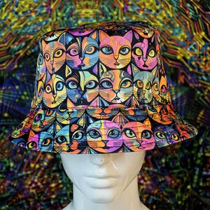 Puede incluir: Un sombrero de pescador colorido con un patrón repetido de caras de gato en varios colores. El sombrero tiene un ala ancha y se muestra en una cabeza de maniquí. El fondo tiene un diseño psicodélico.