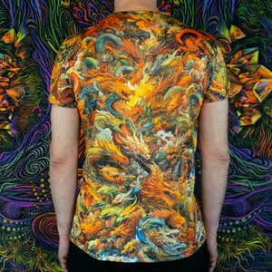 May include: Short-sleeved t-shirt featuring a vibrant, detailed dragon design in shades of orange, yellow, and green. The artwork covers the entire shirt, creating a bold and eye-catching pattern. The person is wearing black pants.