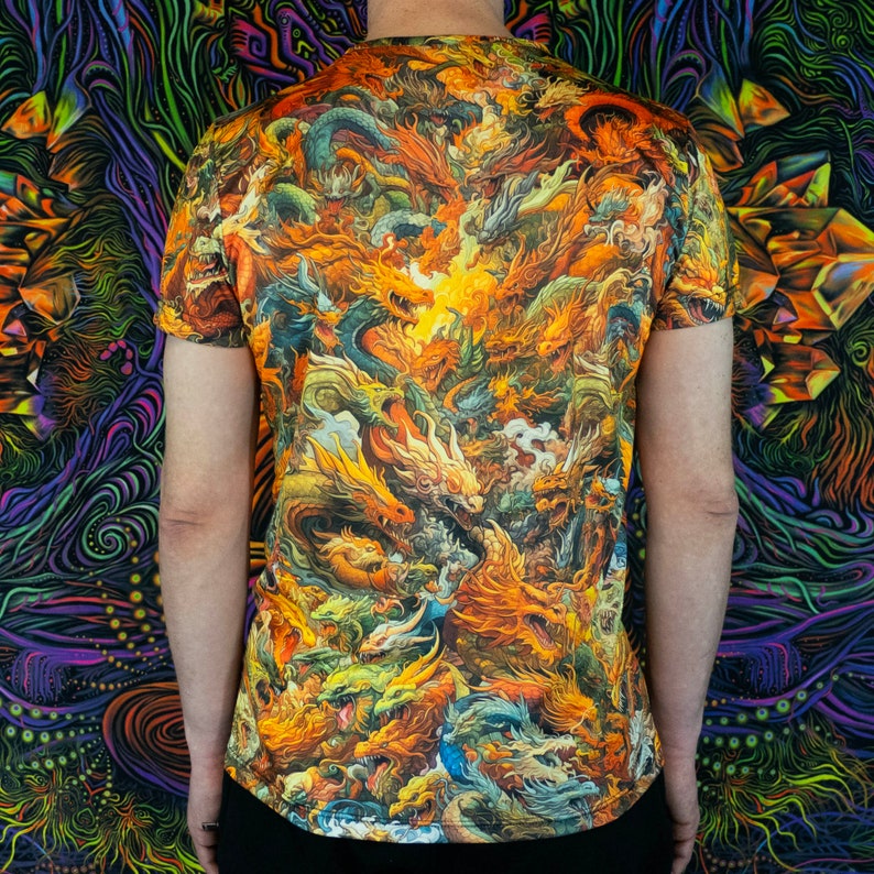 May include: Short-sleeved t-shirt featuring a vibrant, detailed dragon design in shades of orange, yellow, and green. The artwork covers the entire shirt, creating a bold and eye-catching pattern. The person is wearing black pants.