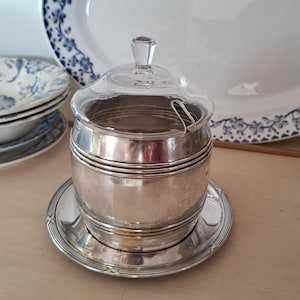 Peut inclure: Un sucrier argenté avec un couvercle en verre transparent et une petite cuillère. Le sucrier repose sur une soucoupe assortie. Des assiettes décoratives avec des motifs floraux bleus sont en arrière-plan.