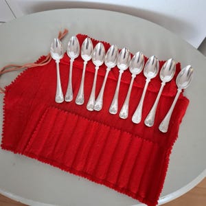 Może przedstawiać: Zestaw dziewięciu srebrnych łyżeczek z ozdobnymi kwiatowymi detalami na uchwytach, ułożonych na jaskrawoczerwonym filcowym wałku na łyżki. Łyżeczki są błyszczące, a wałek jest w głębokim, bogatym odcieniu czerwieni.