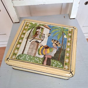 Op de afbeelding: Een vintage blikken doos met een kleurrijke illustratie van een man die gitaar speelt voor een gebouw met een balkon. De scène speelt zich af tegen een blauwe lucht met een maan en sterren. De doos heeft een gele rand met een groen-wit patroon.