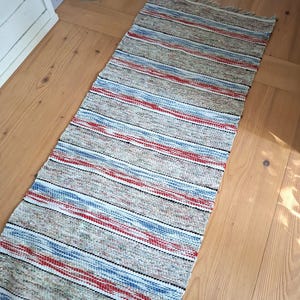 Könnte beinhalten: Ein rechteckiger, gewebter Läufer mit einem Streifenmuster in Beige, Hellblau und Rot. Der Teppich hat eine strukturierte, mehrfarbige Optik mit sichtbaren horizontalen Weblinien und kurzen, ausgefransten Fransen an beiden Enden.