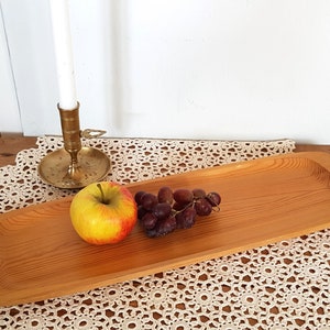 Könnte beinhalten: Ein langes, rechteckiges Holztablett mit natürlicher Holzoptik. Das Tablett liegt auf einer weißen Häkeldeckchen und hat einen roten Apfel und eine Traube darauf.