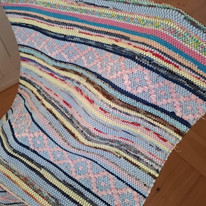 Könnte beinhalten: Ein farbenfroher, gewebter Teppich mit Streifenmuster. Der Teppich weist eine Mischung aus Pastellfarben auf, darunter Rosa, Blau, Gelb und Braun. Das Design beinhaltet ein sich wiederholendes Blumenmuster in der Mitte.