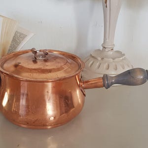 May include: A copper pot with a wooden handle and a lid. The pot has a rounded bottom and a flared lip. The lid has a small metal handle.