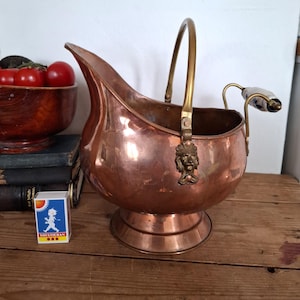 Könnte beinhalten: Ein kupferner Kohleeimer mit Messinggriff und dekorativem Löwenkopf. Der Eimer hat einen Ausguss und einen Keramikgriff an der Seite. Der Eimer steht auf einer Holzoberfläche.