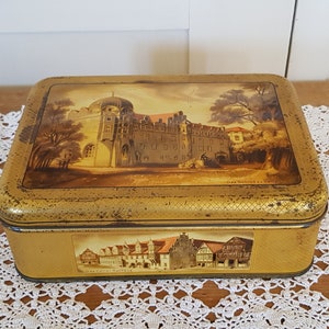 Op de afbeelding: Een vintage gouden metalen blik met een geschilderd beeld van een kasteel en de tekst "Das Schloss zu Celle" op de bovenkant. De onderkant van het blik heeft een geschilderd beeld van een gebouw met de tekst "Das Celler Rathaus".
