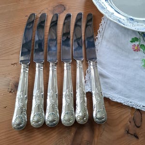 Peut inclure: Un ensemble de six couteaux argentés aux manches ornés. Les couteaux sont disposés sur une surface en bois, avec une assiette décorative et un tissu bordé de dentelle en arrière-plan. Les manches présentent un motif de coquillage et floral.