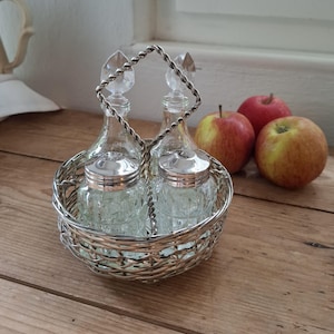 Könnte beinhalten: Ein Vintage-Gewürzset aus Silber mit zwei Glasflaschen und zwei Gläsern mit silbernen Deckeln, in einem geflochtenen Korb. Das Set hat einen gedrehten Griff. Drei Äpfel liegen daneben.