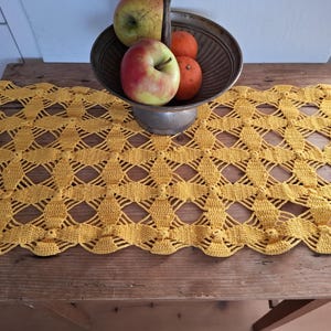 Può includere: Un runner da tavolo in crochet giallo con un motivo a rombi. Il runner è su un tavolo di legno con una ciotola di mele e arance.