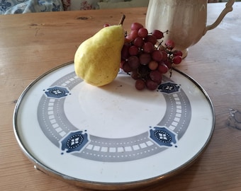 Op de afbeelding: Een ronde serveerschaal met een zilveren rand en een witte kern. De schaal heeft een grijs en blauw geometrisch ontwerp. Een gele peer en een tros rode druiven liggen op de schaal, met een beige kan op de achtergrond.