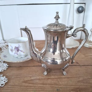 Puede incluir: Una cafetera plateada antigua con un asa, un pico y una tapa decorativos. La cafetera se apoya sobre tres pequeños pies. Una taza y un platillo con un diseño floral están en el fondo. Los artículos están sobre una superficie de madera.