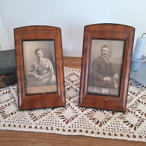 Op de afbeelding: Twee antieke houten fotolijsten met portretten. De lijsten zijn warm bruin van kleur en hebben een gebogen bovenkant. De zwart-wit portretten tonen mensen uit een vervlogen tijdperk. De lijsten staan op een kanten kleedje.