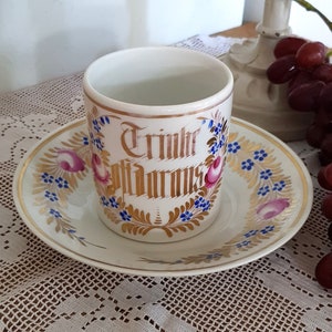 Puede incluir: Un juego de taza y platillo de porcelana blanca con ribetes dorados y diseños florales azules y rosas. La taza tiene las palabras "Trinke Gl"." escritas en letras doradas.