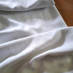 Può includere: Primo piano di tessuto bianco con morbide pieghe e sottili motivi floreali. Il tessuto sembra essere un materiale leggero e traspirante, forse lino o cotone. L'immagine evidenzia la trama e il drappeggio del tessuto.