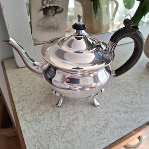 Könnte beinhalten: Eine glänzende silberne Teekanne mit einem dunklen Holzgriff und einem schwarzen Knopf auf dem Deckel. Die Teekanne hat einen gebogenen Ausguss und steht auf drei kleinen Füßen. Die Teekanne spiegelt die Umgebung wider.