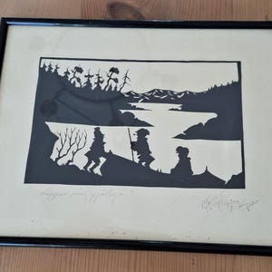 Könnte beinhalten: Gerahmter Schwarz-Weiß-Silhouette-Kunstdruck. Das Kunstwerk zeigt eine Landschaft mit Figuren, Bäumen, Bergen und Wasser. Der Druck enthält Text und eine Signatur. Der Rahmen ist schwarz.