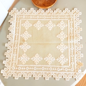 May include: A square, off-white crocheted tablecloth with a delicate, intricate pattern. The tablecloth has a border of small, white squares and a larger, central square with a repeating pattern of white diamonds.