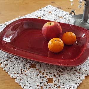 Puede incluir: Una fuente de servir rectangular ligeramente redondeada de cerámica roja. La fuente está llena de fruta, incluyendo una manzana roja y dos mandarinas naranjas.