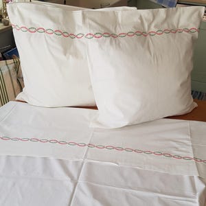 Pode incluir: Lençóis de cama brancos com uma borda bordada em rosa e verde. Os lençóis estão dobrados e dispostos em uma cama.