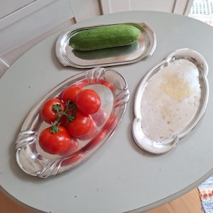 Könnte beinhalten: Drei silberne Serviertabletts, eines mit einer grünen Gurke, eines mit roten Tomaten und eines leer.
