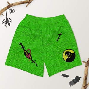Puede incluir: Shorts deportivos verdes con textura de arpillera y un gráfico en blanco y negro de una criatura cosida con un gusano rosa. El gráfico está rodeado por un círculo amarillo. Los pantalones cortos tienen una cruz negra cosida en la pierna derecha y una cruz negra cosida con un gusano rosa en la pierna izquierda.