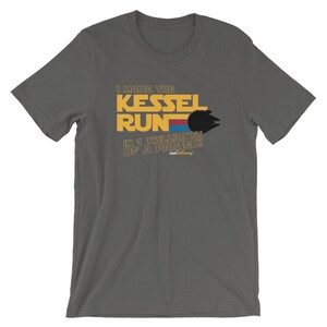 Op de afbeelding: Een donkergrijs T-shirt met een gele en rode afbeelding met de tekst "I made the Kessel Run in 1 trillionth of a parsec!" met een silhouet van de Millennium Falcon.