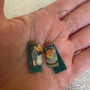 May include: A pair of dangle earrings featuring silver-toned wire hooks and layered rectangular metal charms. The charms display a mix of teal green grid patterns, light blue floral motifs, and small yellow and orange accents on the top pieces.