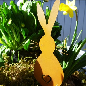 Könnte beinhalten: Ein gelber Metall-Hasen-Ausschnitt mit einer Herzform in der Mitte, umgeben von grünen Pflanzen und gelben Blumen.