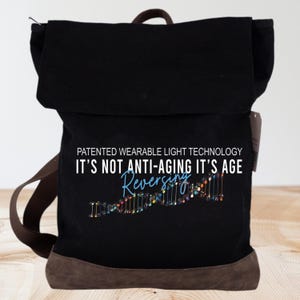 May include: Black canvas backpack with brown leather trim. It features a white graphic with the text "Patented Wearable Light Technology It's Not Anti-Aging It's Age Reversing" and a colorful DNA strand graphic.