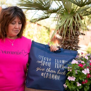 May include: A woman wearing a pink t-shirt with the text "Gramnie" in black script and a heart. She is holding a blue tote bag with the text "Love them, spoil them, give them back #Gramnie" in gold script.