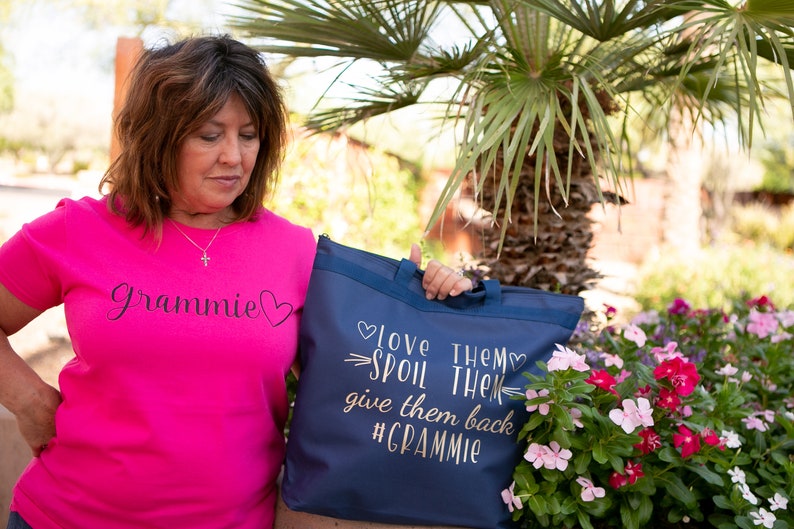 May include: A woman wearing a pink t-shirt with the text "Gramnie" in black script and a heart. She is holding a blue tote bag with the text "Love them, spoil them, give them back #Gramnie" in gold script.