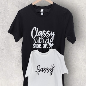 May include: Two t-shirts, one black and one white, with a matching design. The black t-shirt says "Classy with a side of..." with a small heart. The white t-shirt says "Sassy".