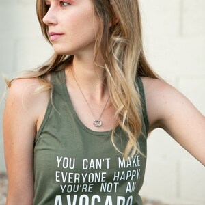 May include: A woman wearing a green tank top with the text "You can't make everyone happy. You're not an avocado." The tank top has a white print. She is wearing black jeans and a silver necklace.