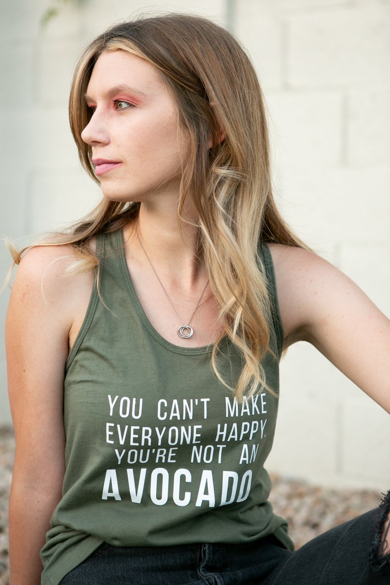 May include: A woman wearing a green tank top with the text "You can't make everyone happy. You're not an avocado." The tank top has a white print. She is wearing black jeans and a silver necklace.