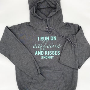 May include: A dark gray hooded sweatshirt with the text "I run on caffeine and kisses #MOMMY" printed in light blue.