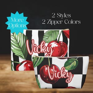 Può includere: Due trousse da toilette a scacchi neri e bianchi con un design a ciliegie e foglie. Le trousse sono personalizzate con il nome "Vicky".
