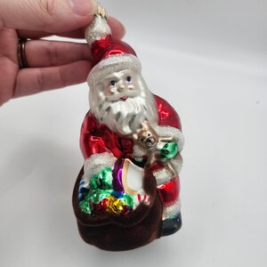 May include: A Christmas ornament shaped like Santa Claus, featuring a red suit with white trim and a matching hat. Santa holds a brown sack filled with colorful gifts and a small teddy bear. The ornament has a shiny, reflective surface.