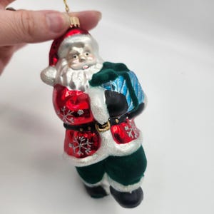 May include: A Christmas ornament in the shape of Santa Claus. The ornament features a red coat with white trim, a green velvet pants, and a blue sack. The ornament is detailed with snowflakes and a gold belt.