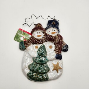 May include: A ceramic snowman ornament with two snowmen figures. The snowmen wear brown hats and scarves, with a green Christmas tree and a gold star. The ornament has a tag that says "WORKBENCH".
