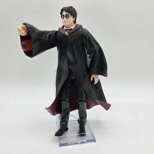 May include: A Harry Potter action figure, wearing a black robe with burgundy lining, a white shirt, and round glasses. The figure is posed with an outstretched arm, as if casting a spell. The figure is standing on a clear plastic stand.