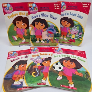 May include: A collection of six children's books featuring Dora the Explorer. The books have red and yellow covers with illustrations of Dora and her friends. Titles include "Explore With Dora," "Dora's Show Time!," and "Dora Saves the Game."