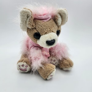 May include: A plush brown bear with a cream-colored muzzle and ears, wearing a pink feathered boa and matching hat. The bear has large black eyes and pink paw pads, creating a whimsical and charming aesthetic.