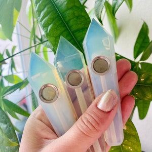 May include: Three iridescent opal crystal pipes with metal bowls. The pipes are held in a hand and are pointed upwards.
