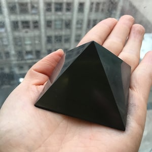 May include: A black obsidian pyramid crystal, a type of gemstone, resting in the palm of a hand.