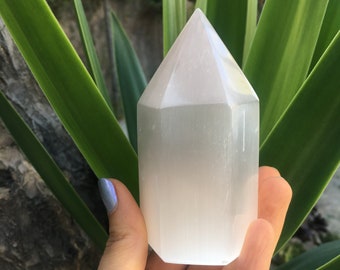 May include: A white selenite crystal tower with a smooth, polished surface. The crystal is shaped like a pyramid with a pointed top and a rectangular base.
