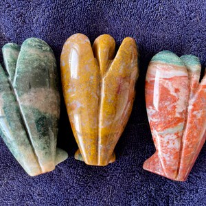 May include: Three carved stone angel figurines. The angel on the left is green, white, and brown. The angel in the middle is yellow, white, and brown. The angel on the right is red, white, and green.