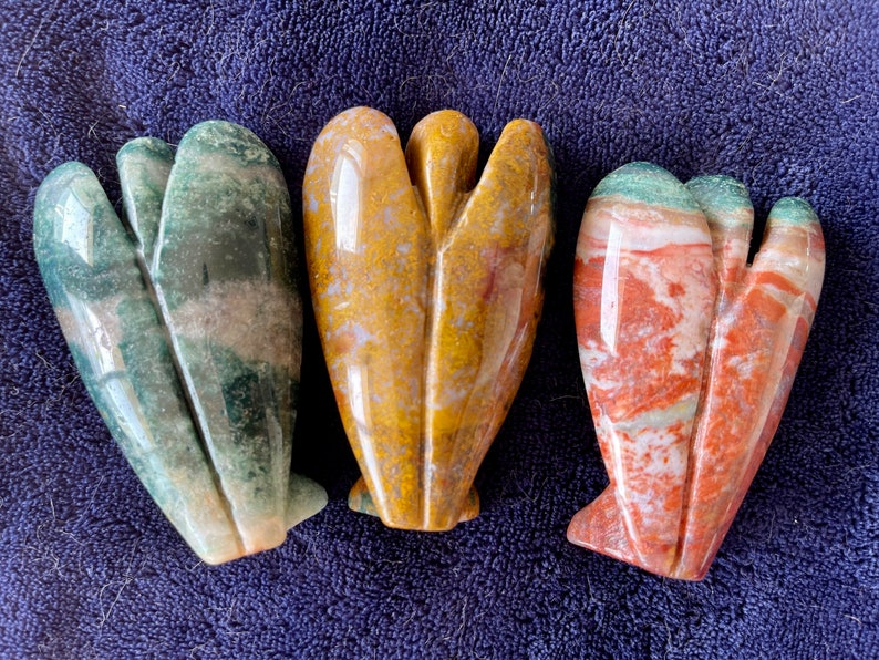 May include: Three carved stone angel figurines. The angel on the left is green, white, and brown. The angel in the middle is yellow, white, and brown. The angel on the right is red, white, and green.